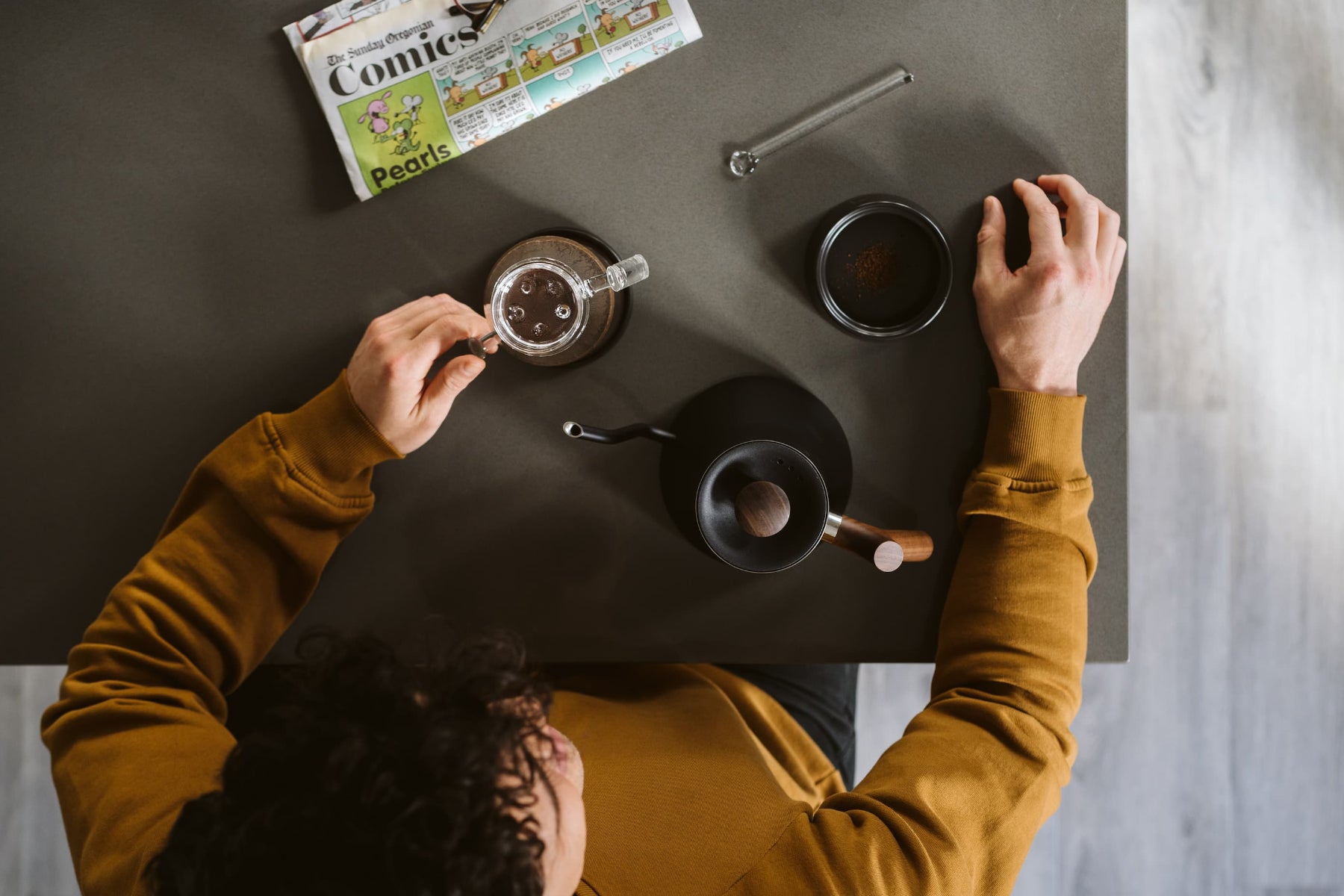 Person making coffee at a table with a magazine and a spoon.