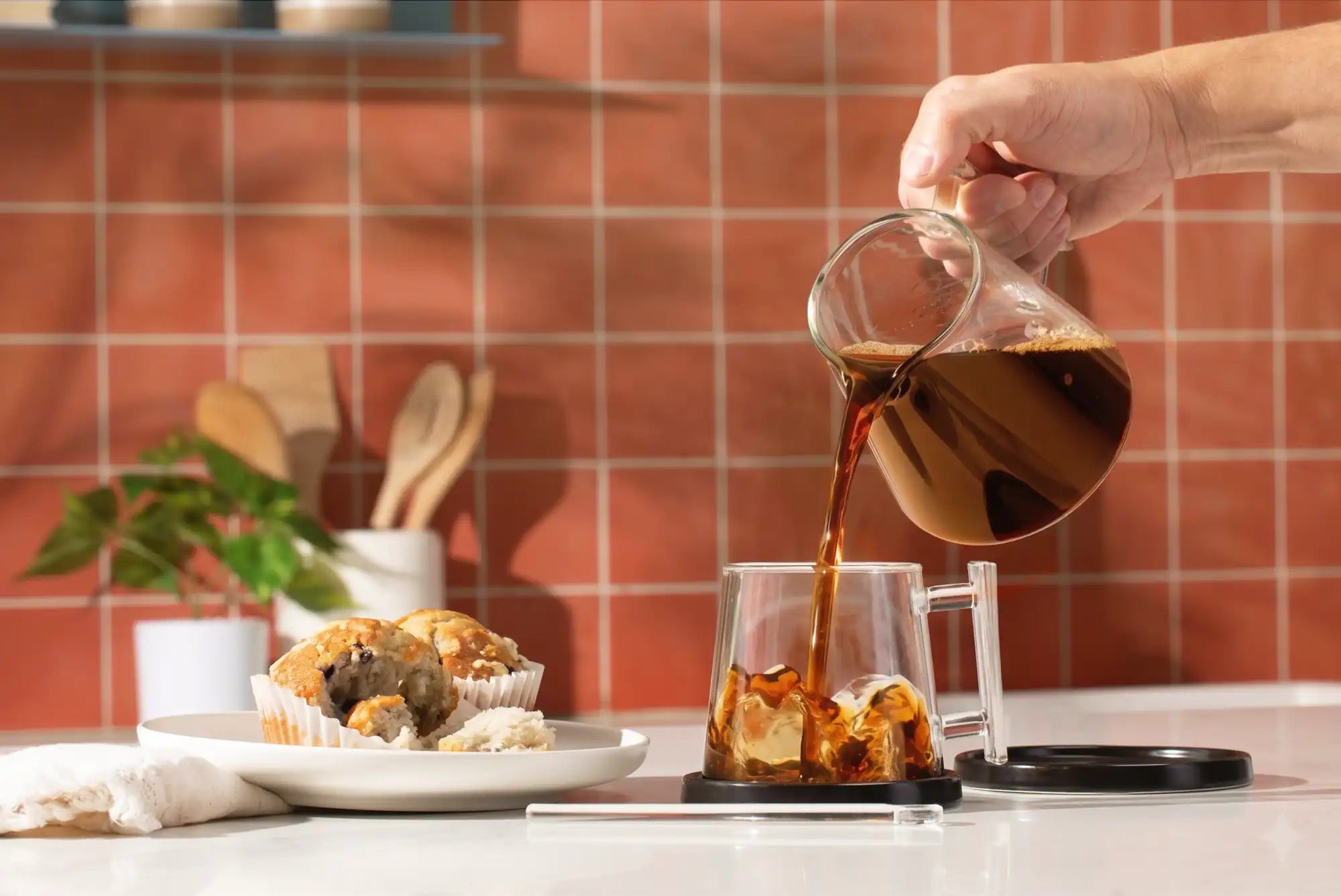 Person pouring coffee into a glass mug with ice and a muffin on a kitchen counter.