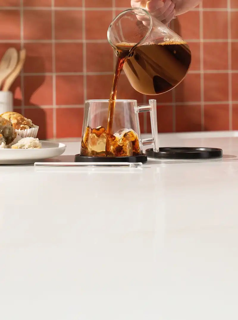 Person pouring coffee from a carafe into a glass of iced coffee on a kitchen counter.